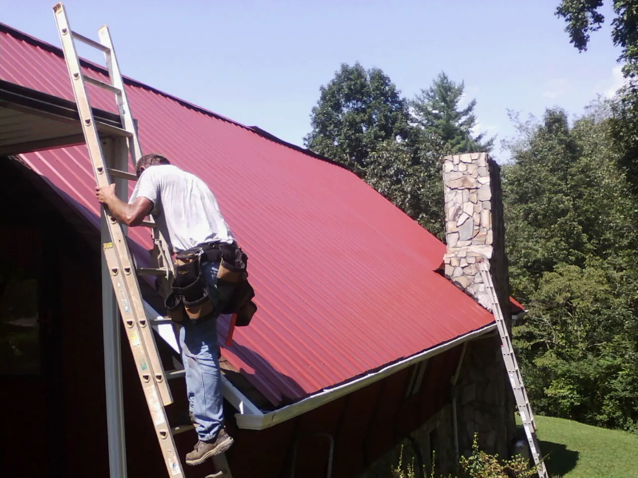 Roofer on a ladder beside a red metal roof and stone chimney — Soffit & Fascia Repair by Roofing Brothers in Fairborn
